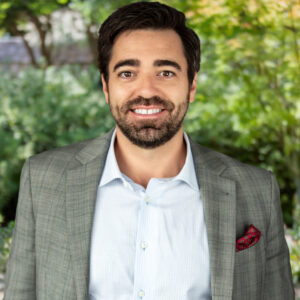 Aaron Luque smiles and wears a white shirt and grey suit jacket against a backdrop of greenery.