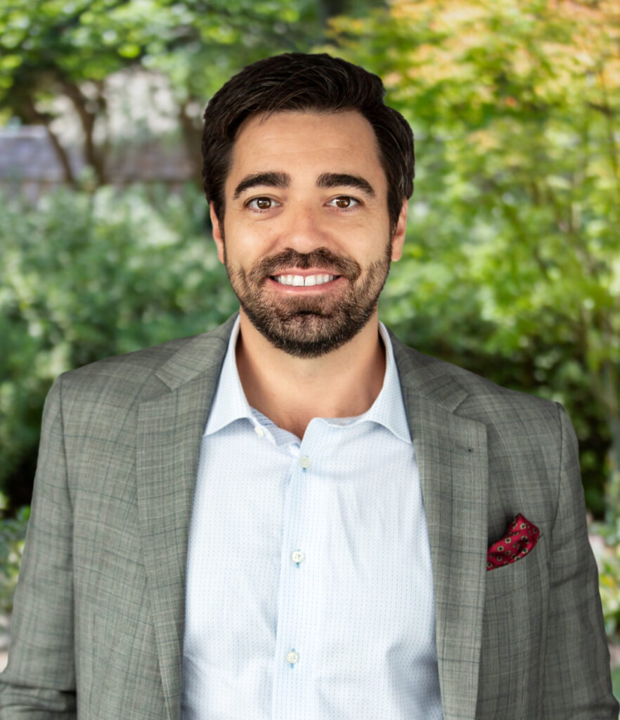 Aaron Luque smiles and wears a white shirt and grey suit jacket against a backdrop of greenery.